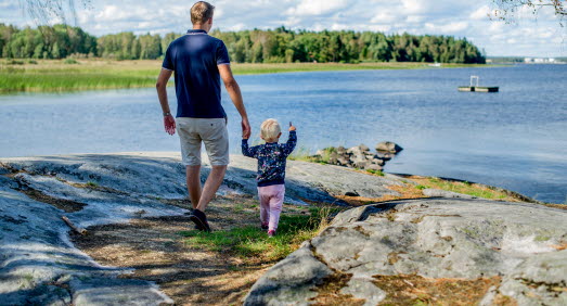 Pappa och barn håller varandra i handen och blickar ut över vattnet. Barnet pekar ut mot vattnet. 