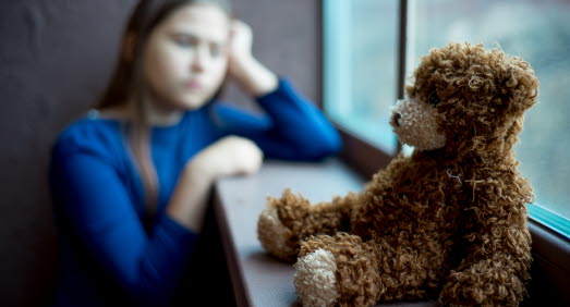 En flicka tittar ut genom ett fönster. I förgrunden sitter en nallebjörn.