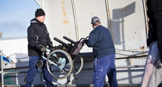 Två män i arbetskläder som kastar en uttjänt rullstol i en återvinningscontainer en solig dag