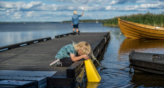 Två barn står på en brygga. Ena barnet fiskar och det andra barnet tittar ner i en vattenkikare. 