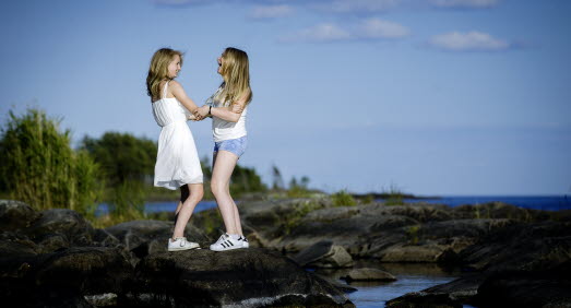 Två tjejer står på en klippa vid vattnet och skrattar och håller varandras händer