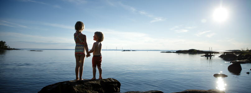 Två barn står på en klippa vid vattnet och håller varandra i handen 