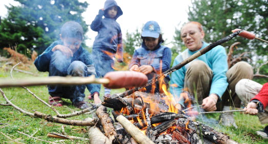 Barn och vuxna sitter vid eld och grillar korv.