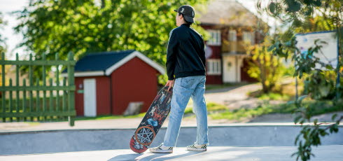 Pojke står med ryggen mot kameran, i skateparken, och håller i en skateboard. 