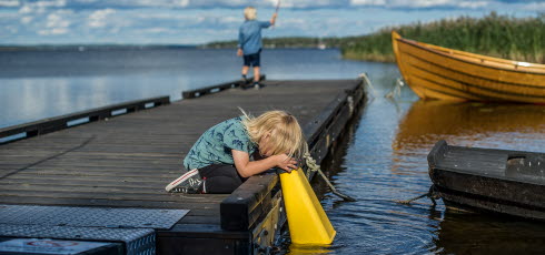 Två barn står på en brygga. Ena barnet fiskar och det andra barnet tittar ner i en vattenkikare. 