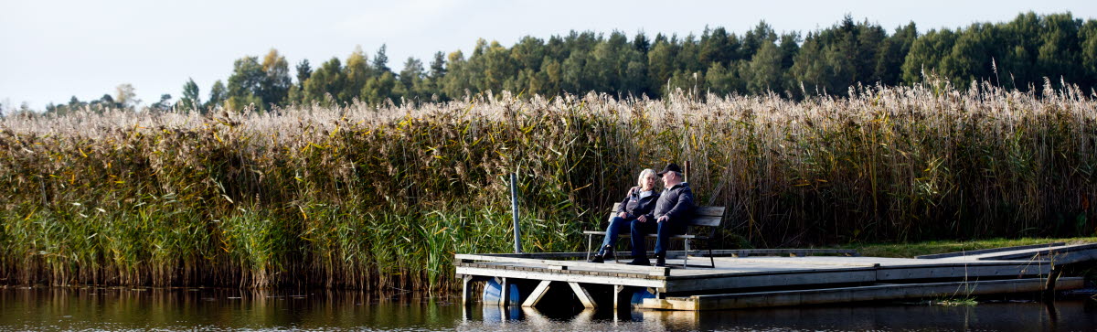 Äldre man och kvinna sitter på en bänk ute på en brygga vid vattnet och pratar med varandra 